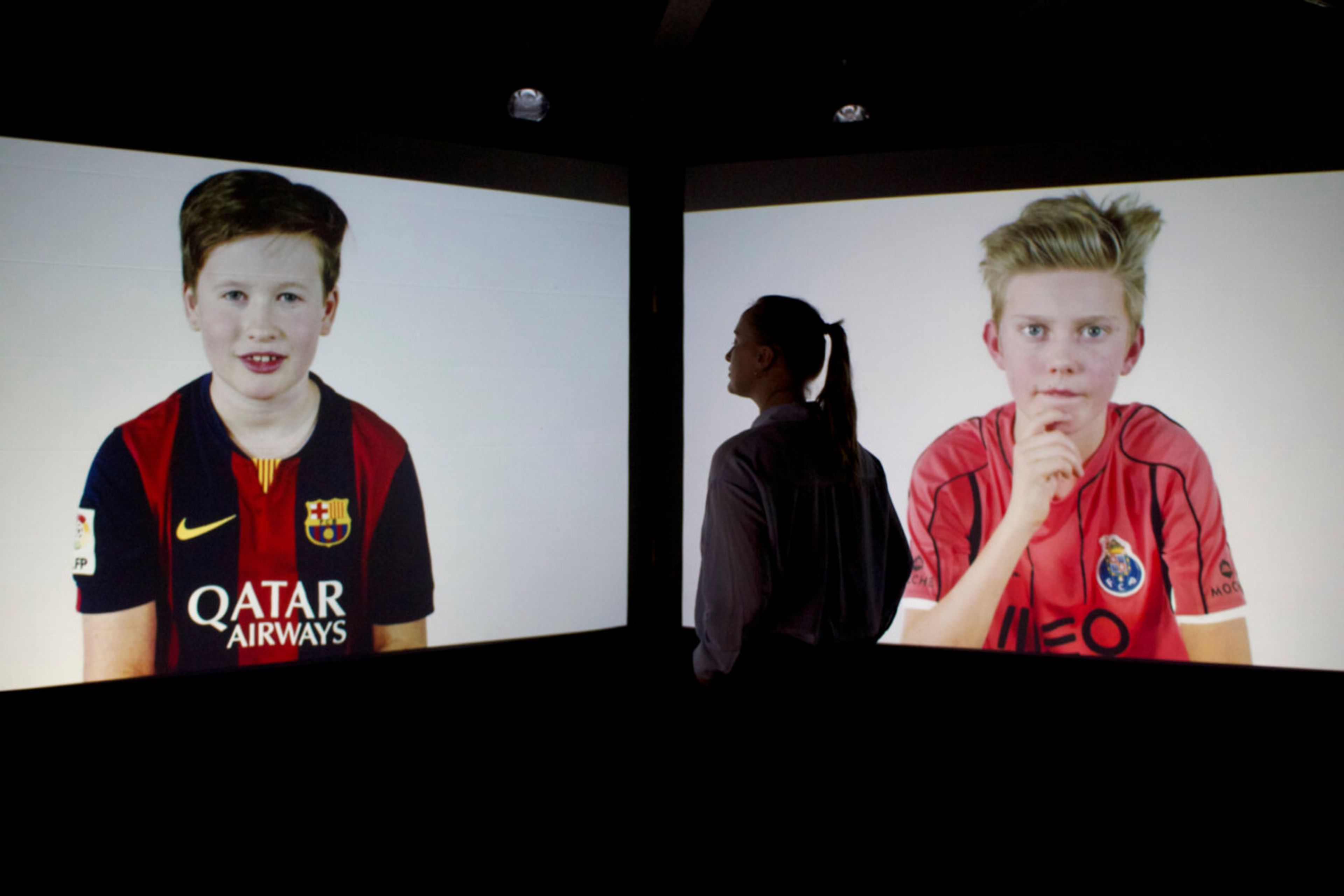 A visitor is watching a 2 channel video art presenting 2 young boys in football strips.