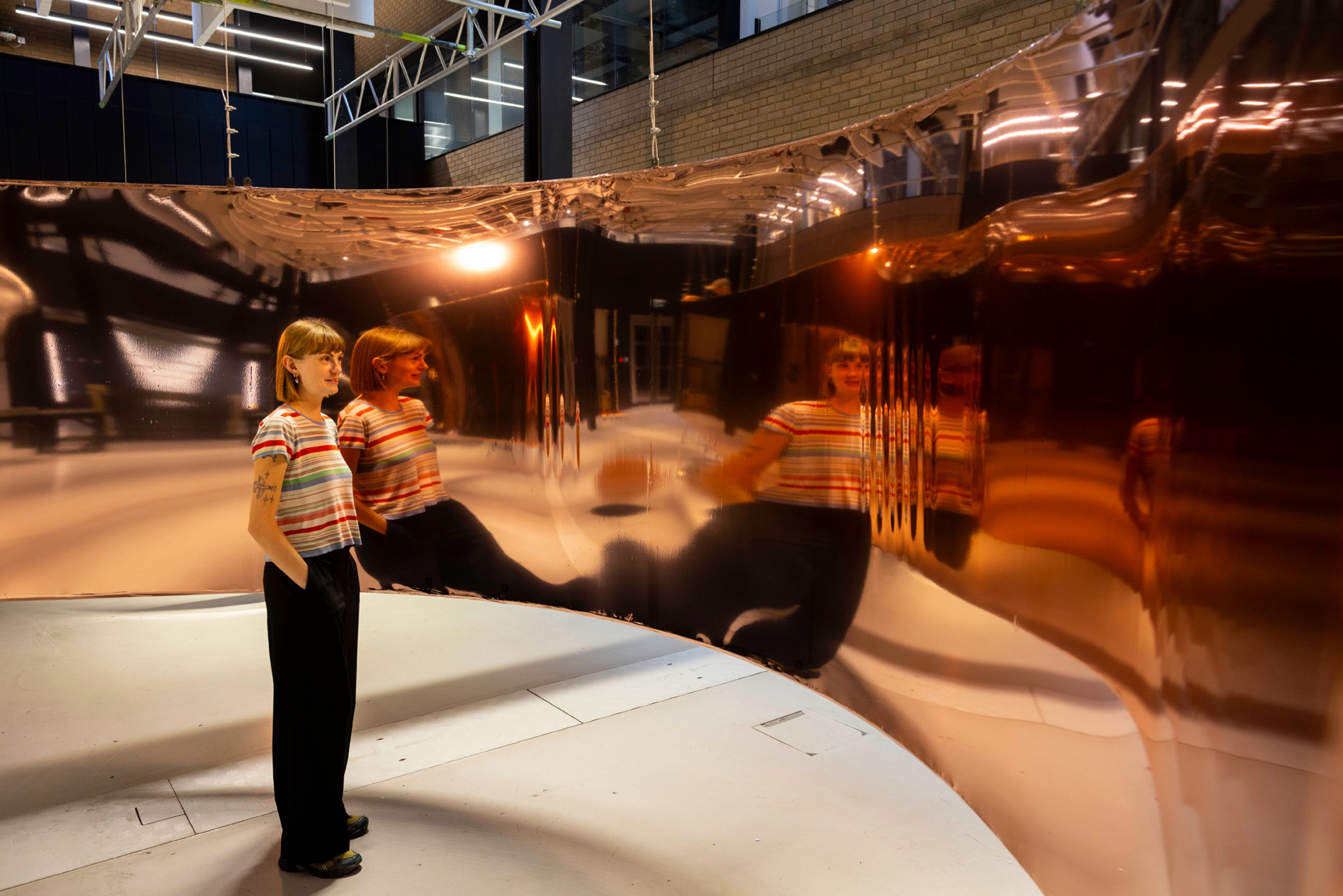 A person stands in front of a reflective copper material that reflects and distorts their image.