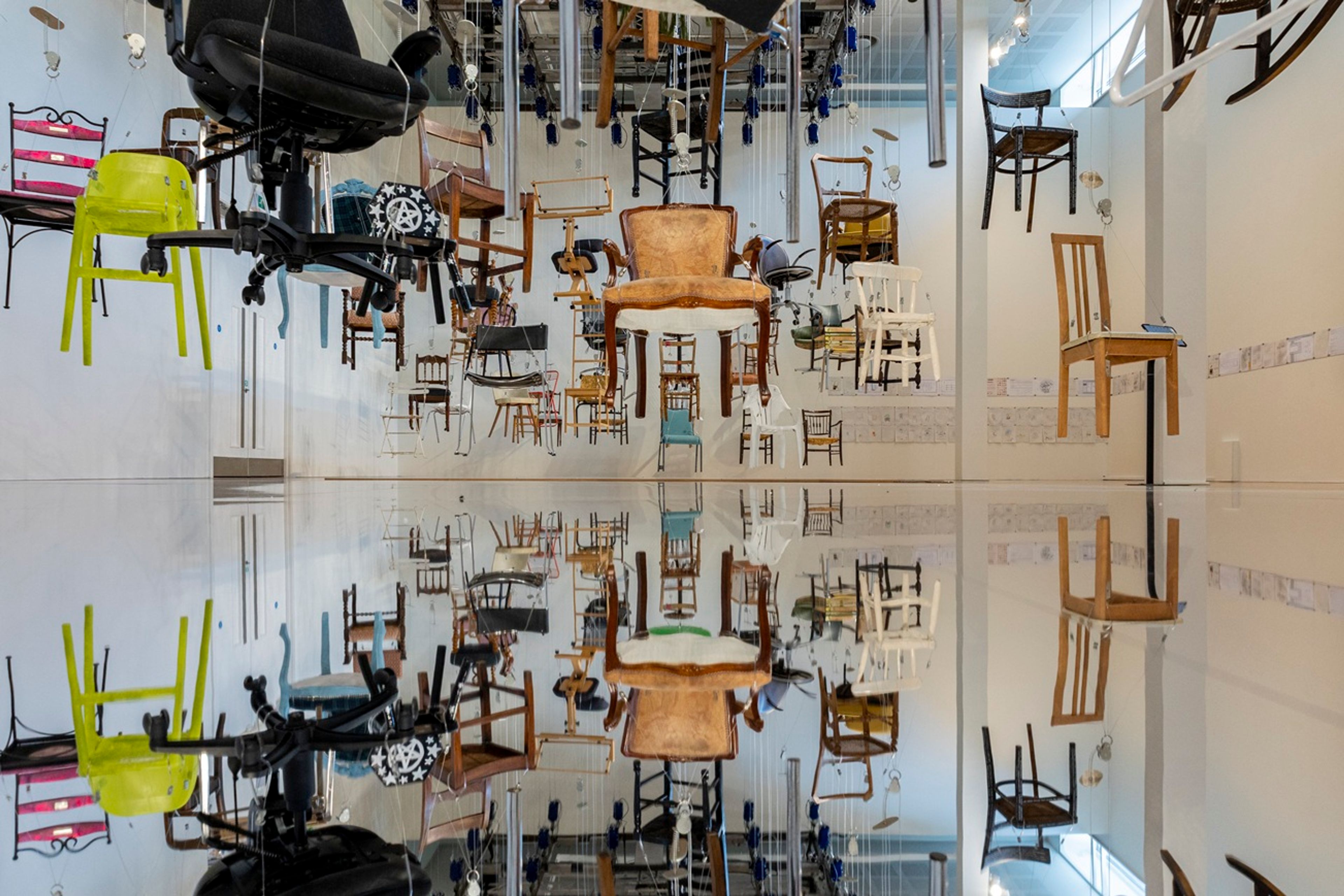 Large gallery installation featuring dozens of suspended chairs hanging at different heights above a reflective floor.