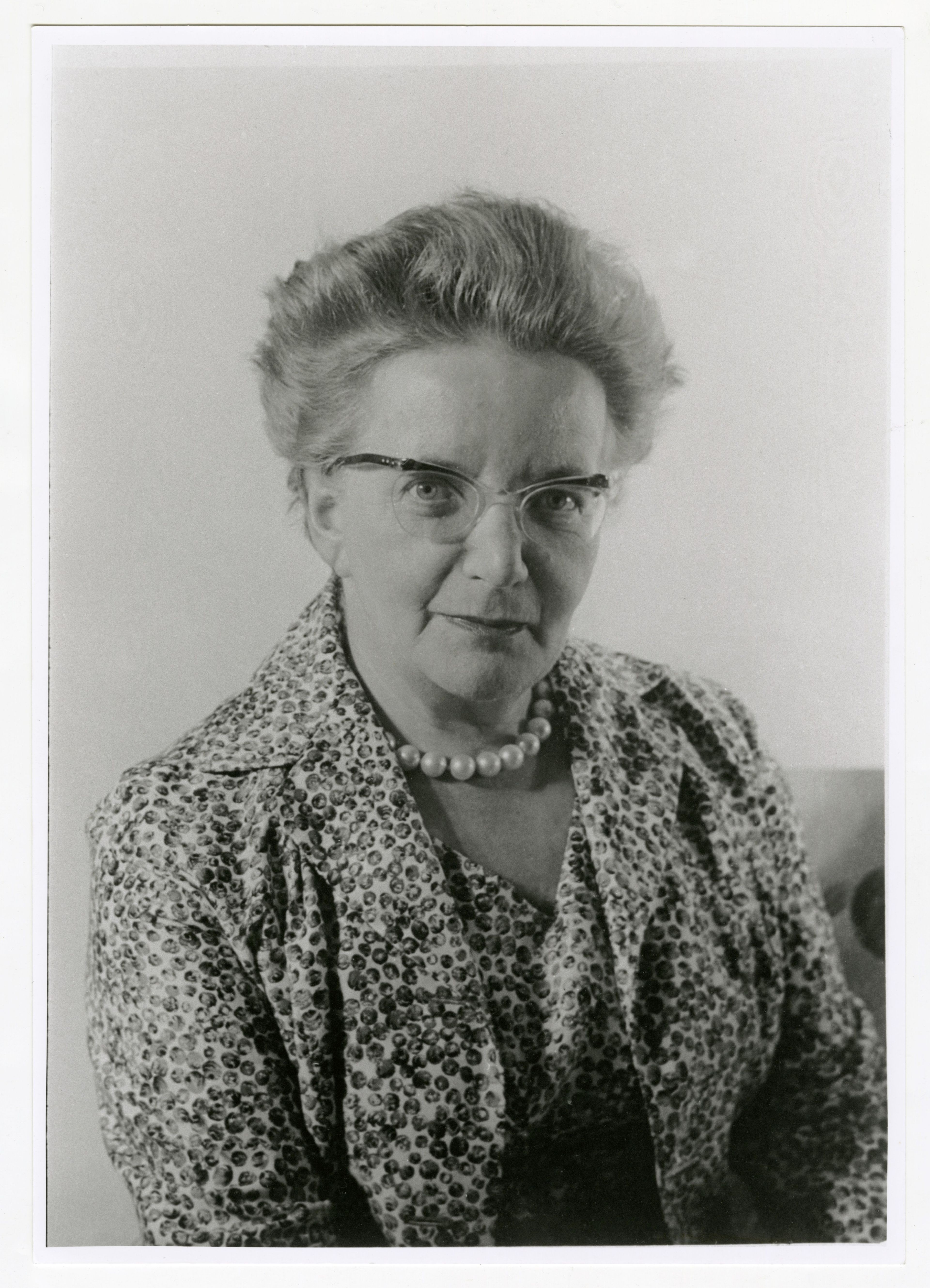 Black and white portrait of Lisel Haas. She is wearing glasses and has short hair. She is wearing a patterned jacket and a pearl necklace.