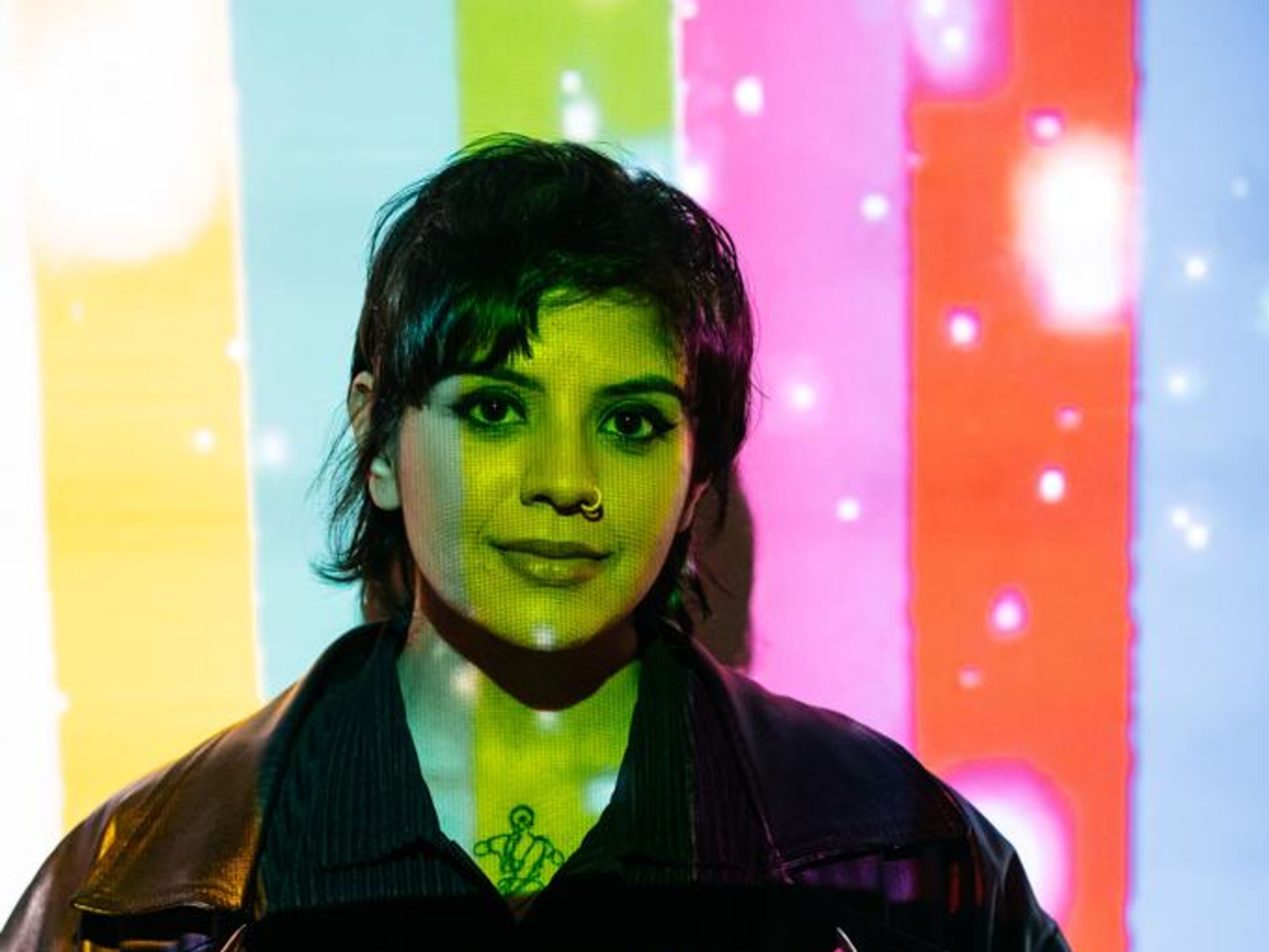 Seema is stood in front of a wall with a multi-coloured projection across their face and the wall.