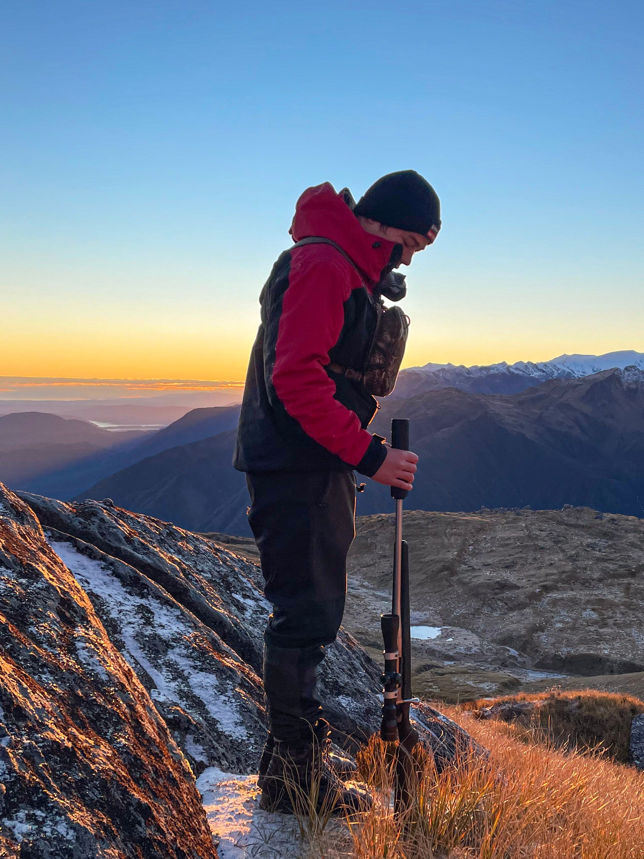 Bayley standing on frosted alpine tops at sunrise with a suppressed bolt-action rifle, snow-capped ranges behind