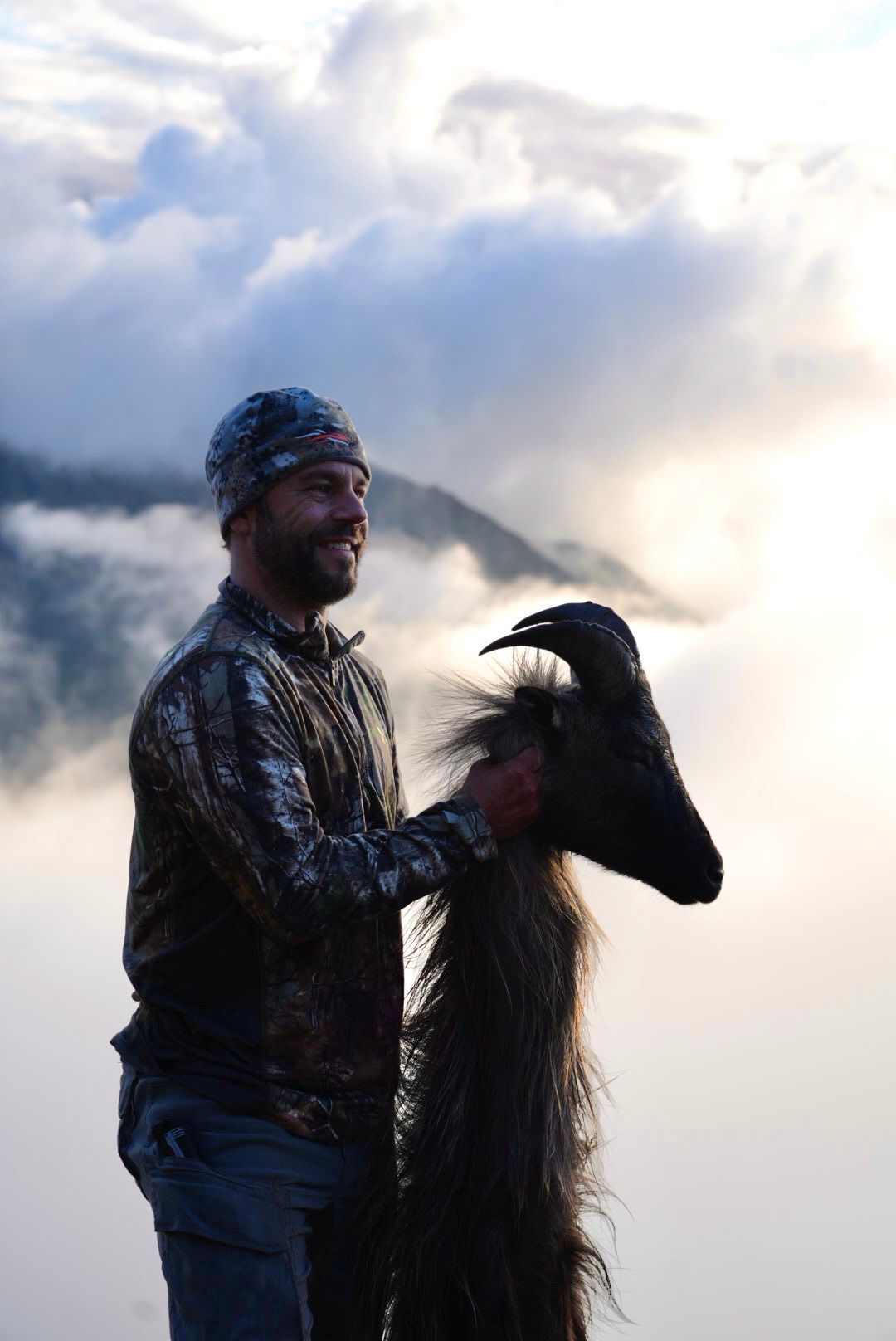 Hunter holding a bull tahr cape above the cloud line at sunrise in the Southern Alps, New Zealand