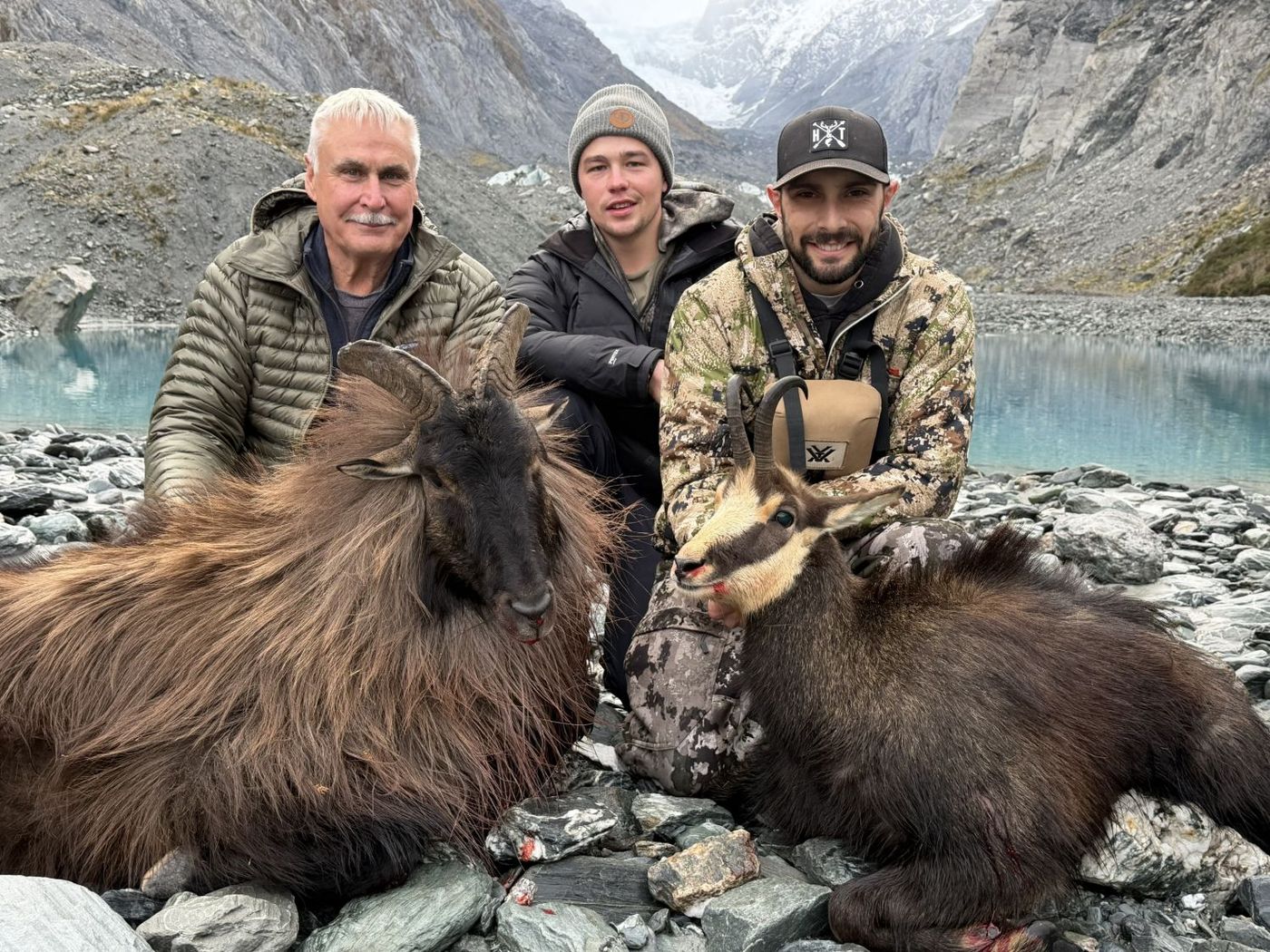 Bayley and two hunters kneel beside a bull tahr and a chamois on a riverbed below a glacier, with a turquoise lake behind.