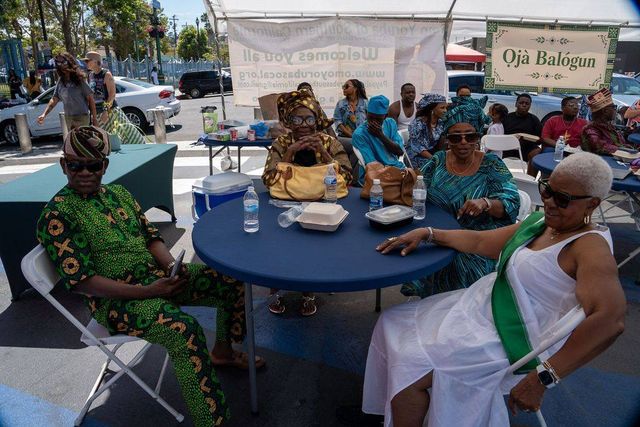 Elders in aṣọ òkè and lace rest at a round table under the Ọjà Balógun sign