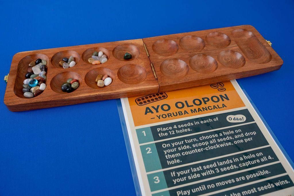 A wooden ayo board with stones beside the Ayo Olopon rules card