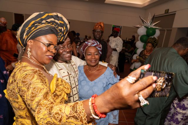 A woman in a gold lace dress and a striped gèlè takes a selfie with two friends on the dance floor
