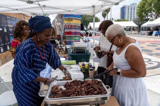 A vendor in indigo àdìrẹ serves suya to a guest in white