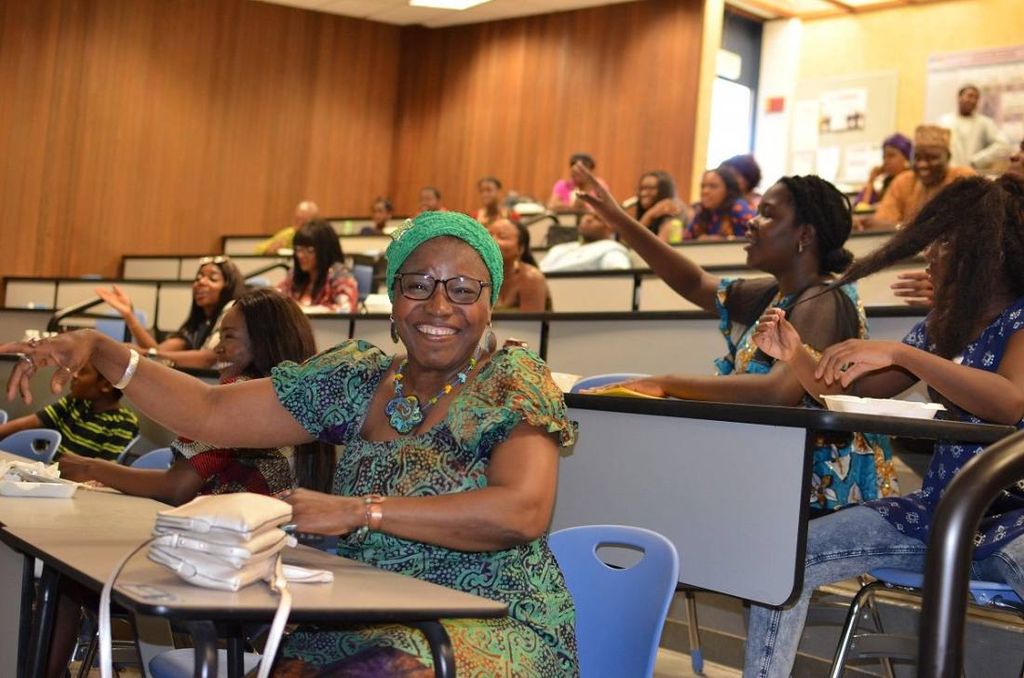 A woman in green print and gèlè laughs with her arms out as the room sings