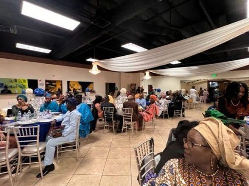 Guests in aṣọ òkè and gèlè seated at round tables under white drapes and chandeliers