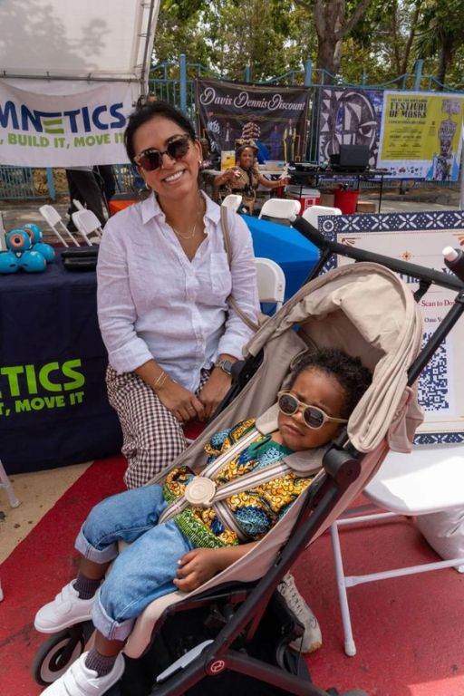 A mother crouches beside her toddler, who wears sunglasses in his stroller, at the STEM tent