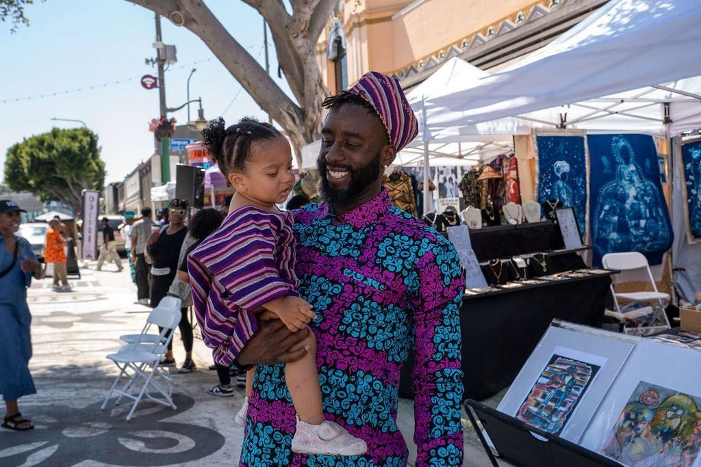 A father in a purple and teal print holds his young daughter beside the art stalls