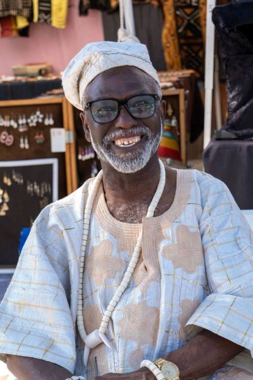 An elder in a pale aṣọ òkè agbada and cap smiles at a vendor stall
