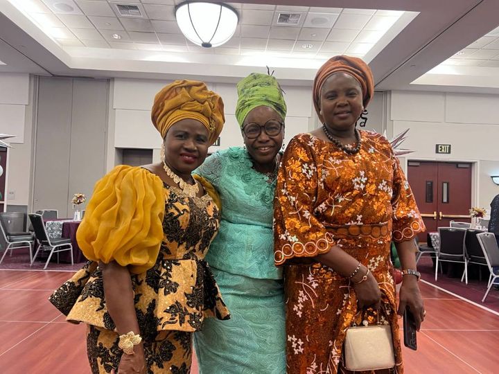 Three women in gold, green and copper gèlè and lace stand together in the hall