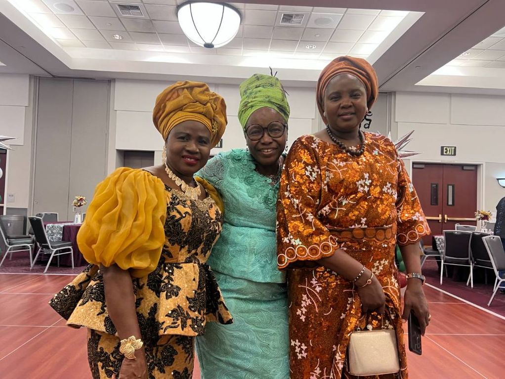 Three women in gold, green and copper gèlè and lace stand together in the hall