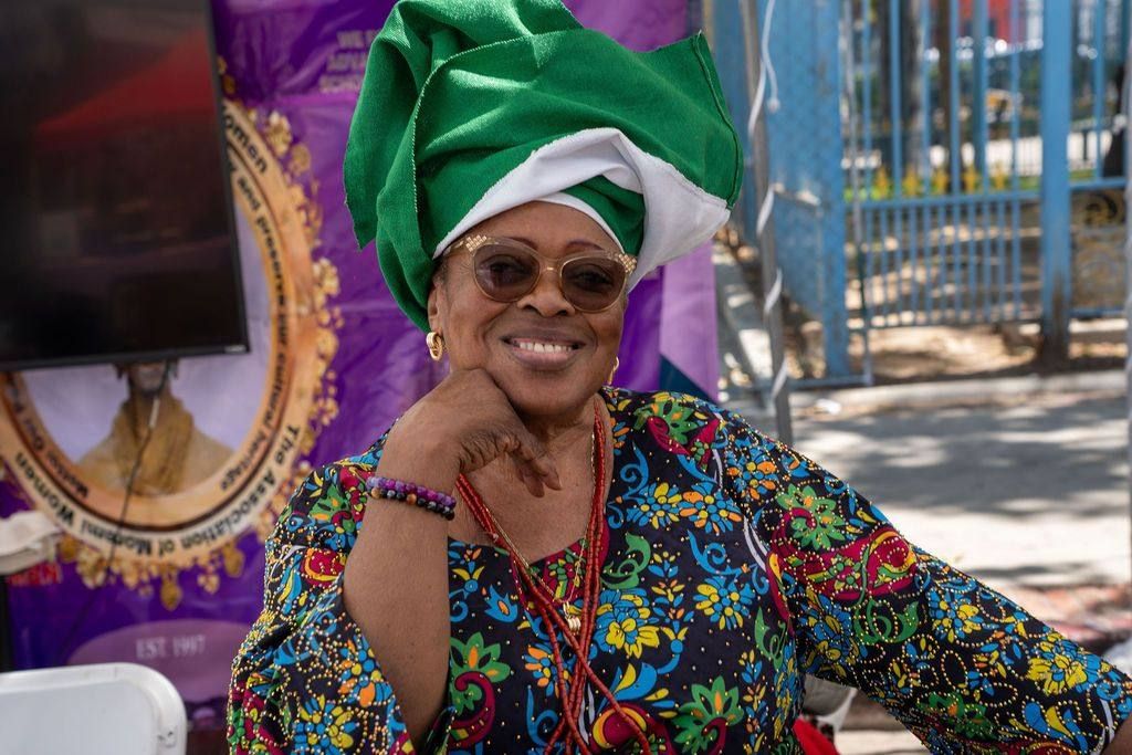 An elder in a green and white gèlè and sunglasses smiles at the market