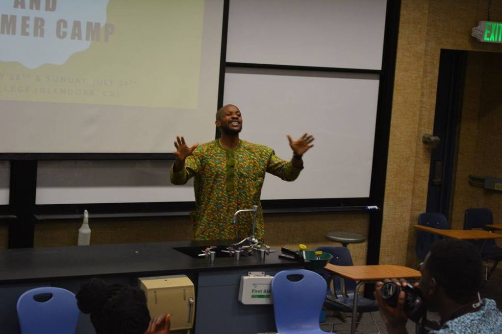 A teacher in green print throws his hands up in front of the summer camp slide