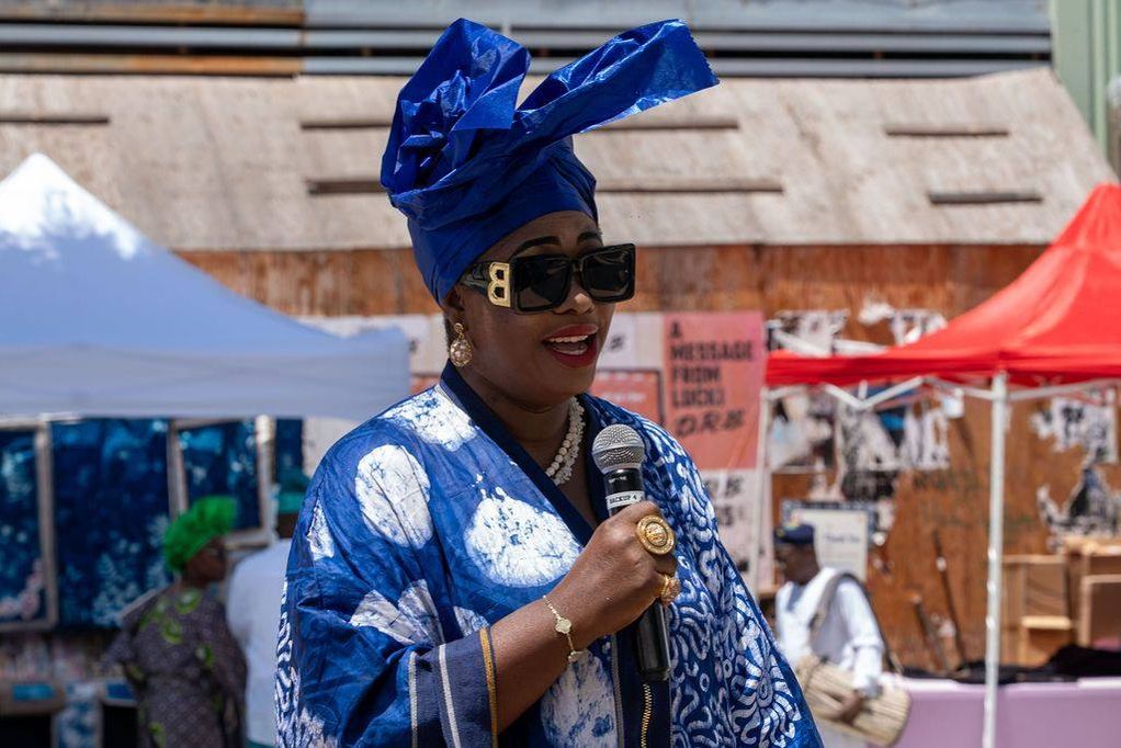 A woman in blue àdìrẹ and a blue gèlè speaks into a microphone