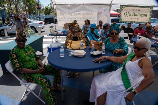 Elders in aṣọ òkè and lace rest at a round table under the Ọjà Balógun sign