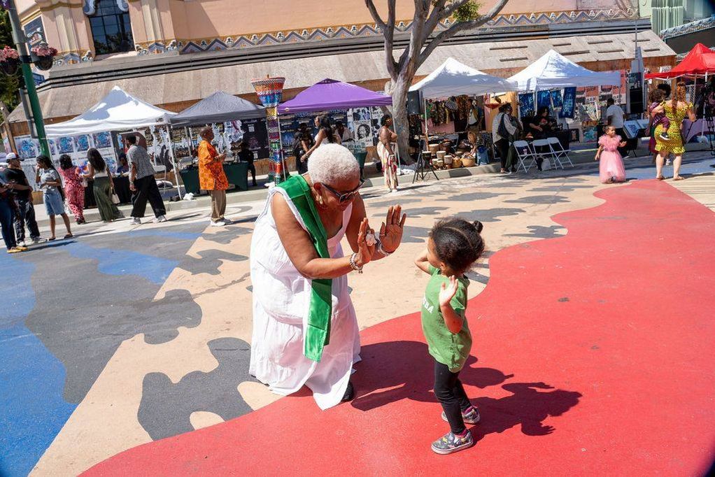 An elder in white with a green sash high-fives a toddler on the painted plaza