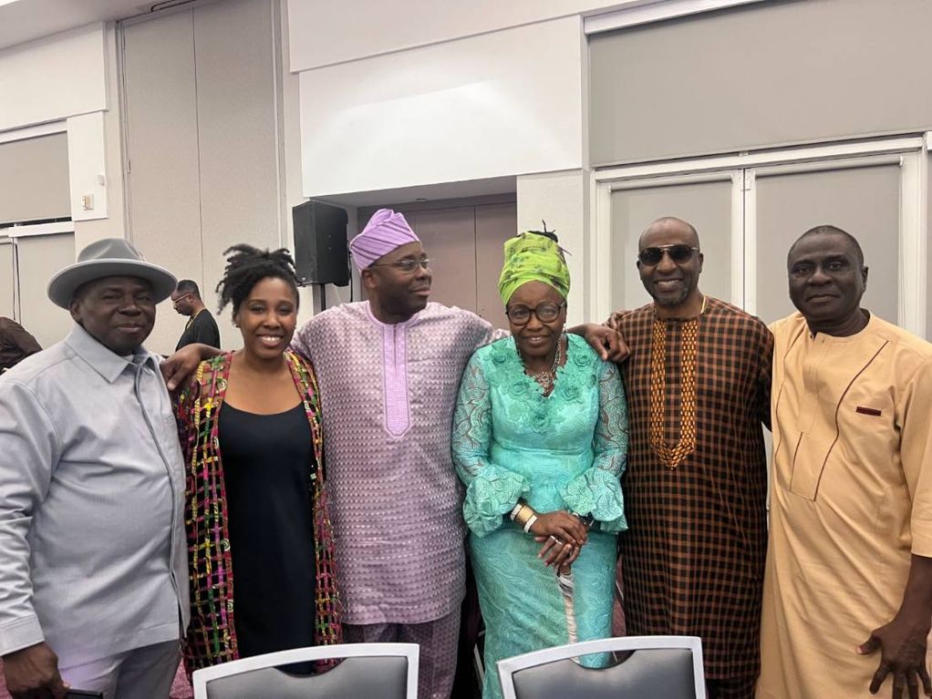 Six guests in agbada, gèlè and a wide-brimmed hat stand arm in arm