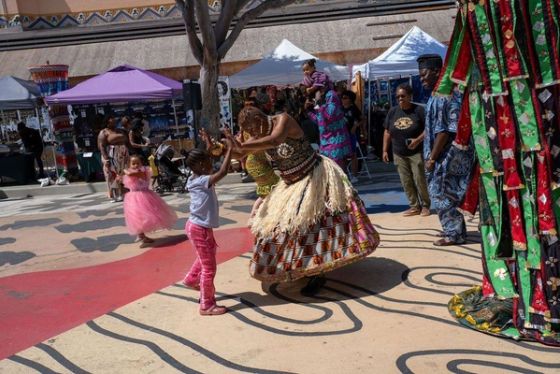 A dancer in a raffia skirt plays with a young girl, the masquerade beside them