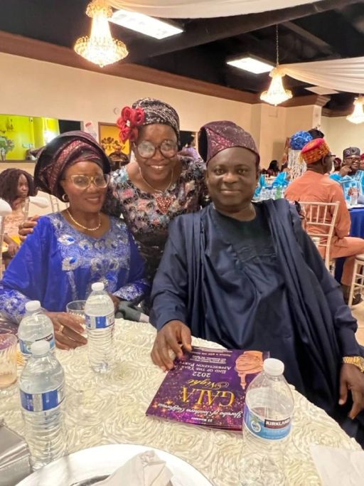 Two women and a man in dark aṣọ òkè smile at their table, a gala programme in hand