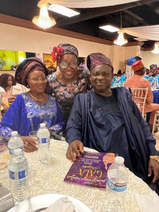 Two women and a man in dark aṣọ òkè smile at their table, a gala programme in hand