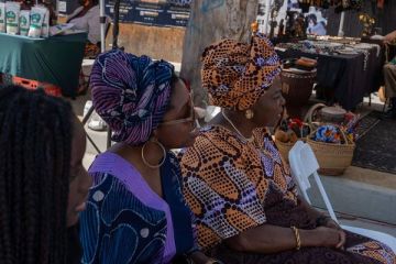 Two women in gèlè listen to the Yoruba lesson beside the market stalls