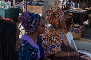 Two women in gèlè listen to the Yoruba lesson beside the market stalls