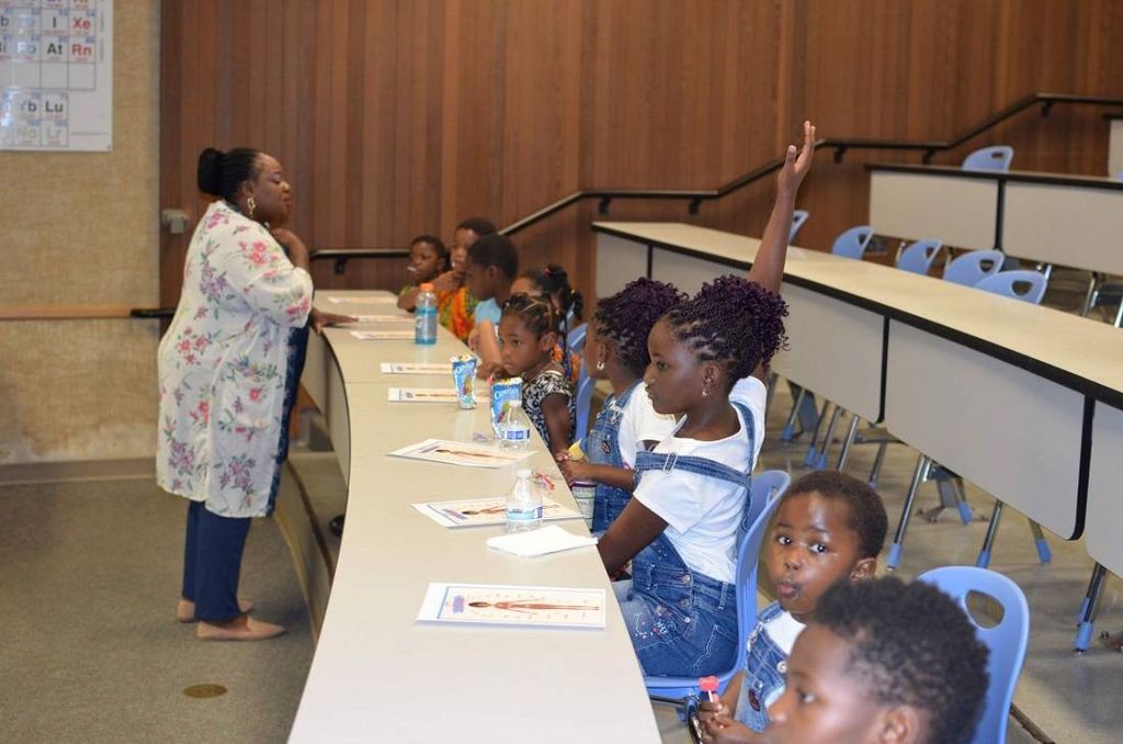 A teacher leads a class of young children in a lecture hall; one girl raises her hand