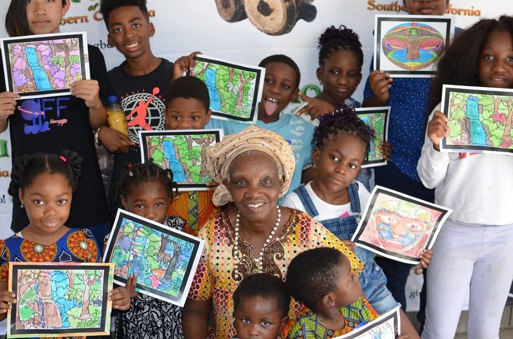 An elder in gèlè sits among a dozen children holding up their coloured drawings in front of the banner