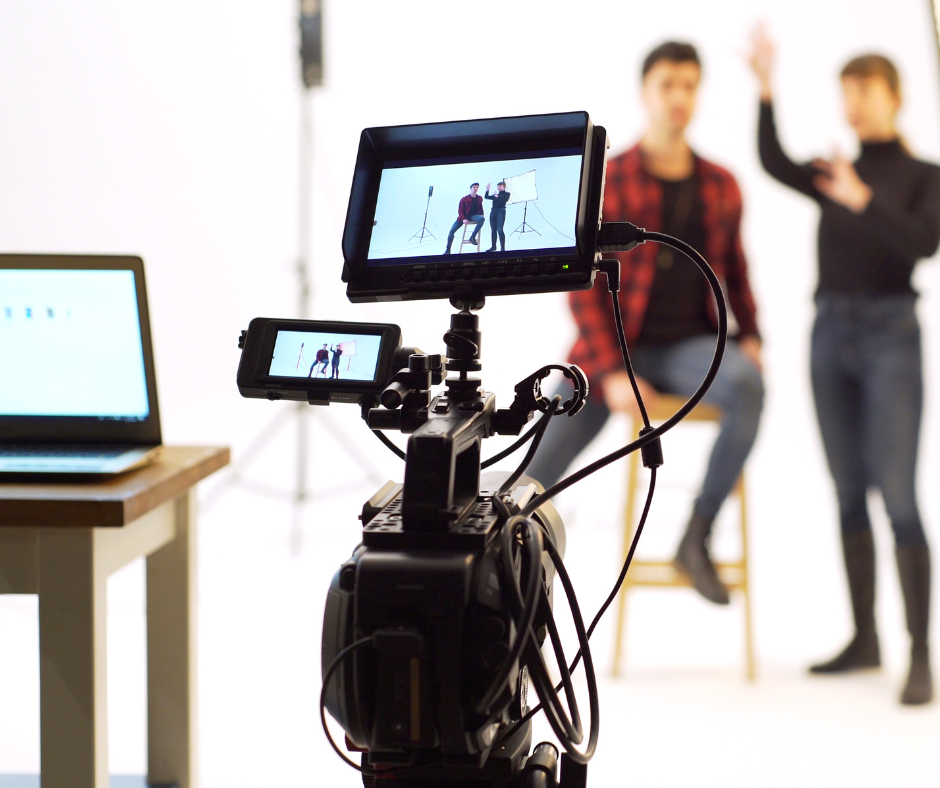 Two people filming in a studio, a camera with a viewing screen is in the foreground with the people in the background in front of a white backdrop