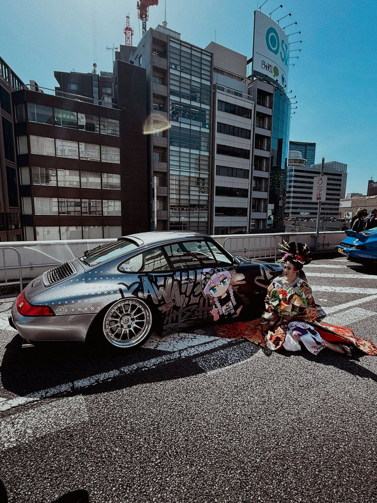 Silver Porsche 964 with anime graffiti livery and woman in traditional kimono sitting next to it on the expressway