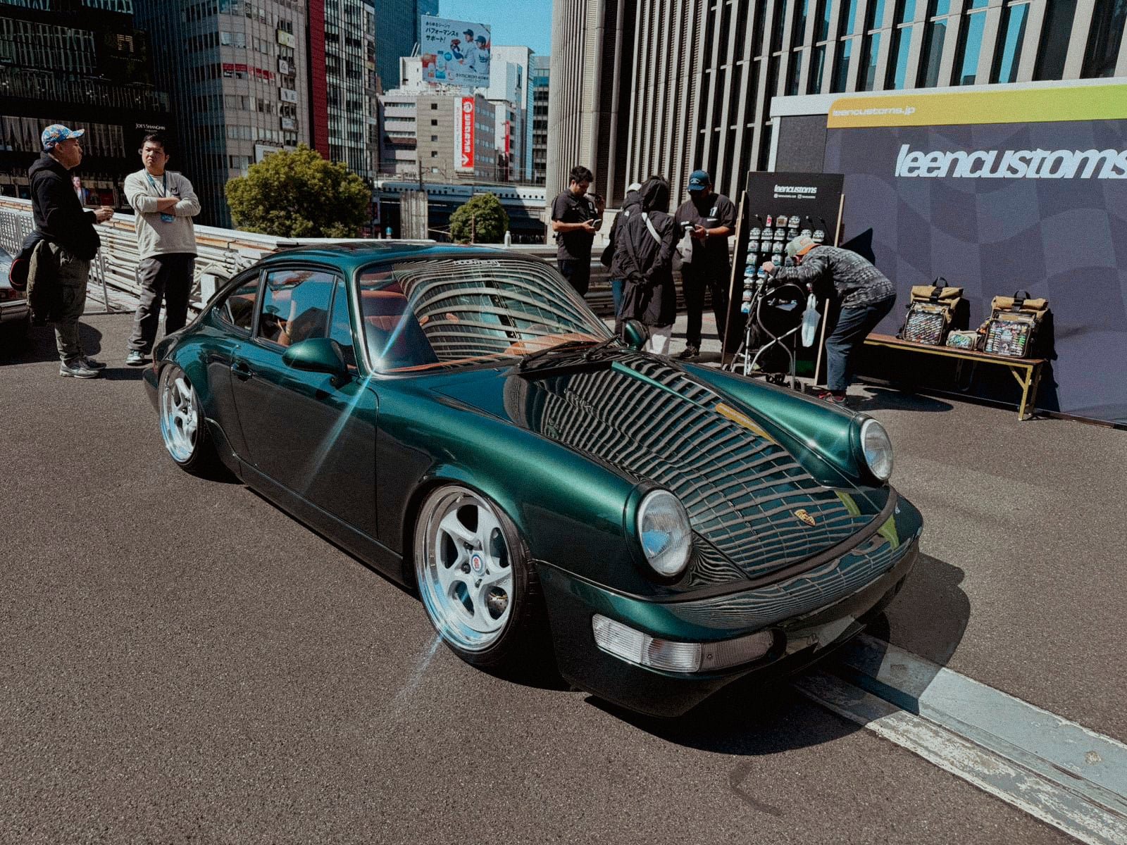 Dark green lowered Porsche 964 with Keen Customs stand behind in Ginza sunlight