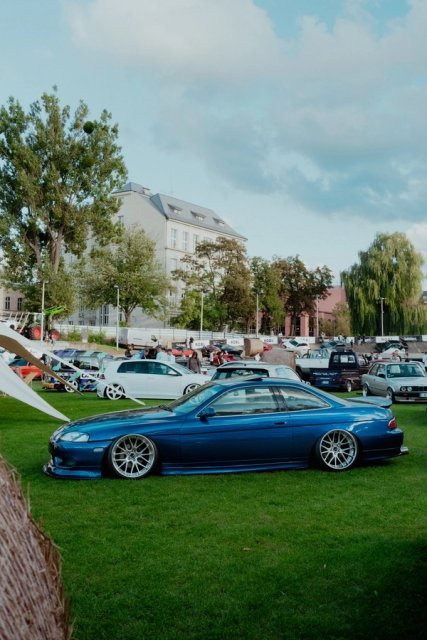 A dark blue BMW E36 coupe on the grass infield, event tents and parked cars behind it