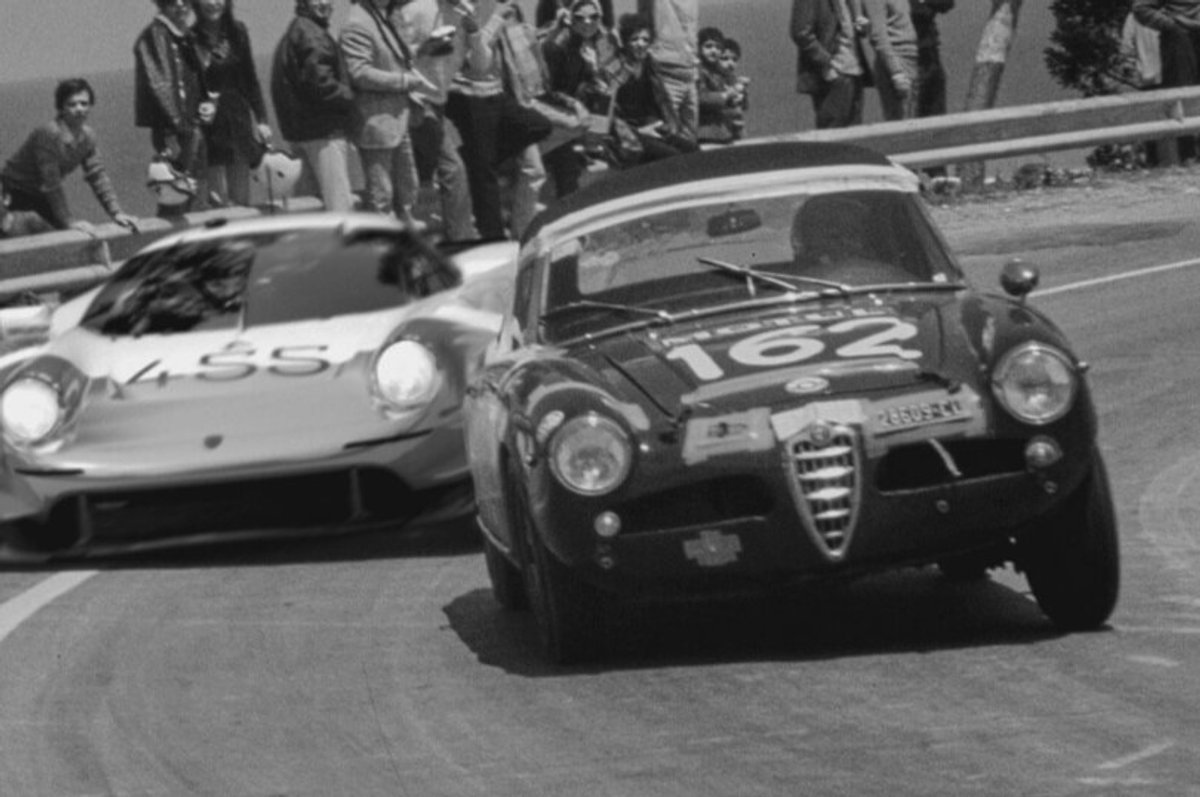 Black-and-white archive photo of an Alfa Romeo with number 162 leading a low white prototype with number 455 through a corner, spectators lining the barrier behind them