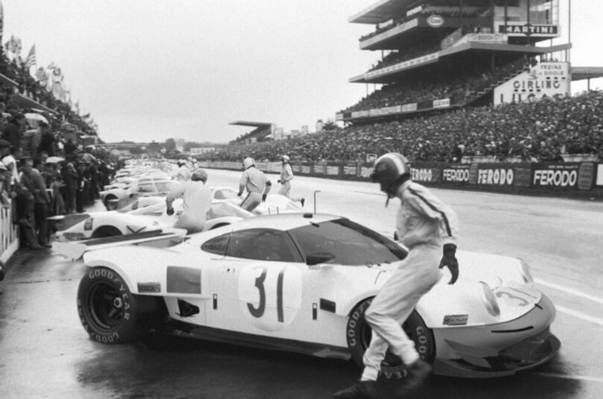 Black-and-white archive photo of a driver in white overalls at the door of a white No. 31 prototype in a wet pit lane, full grandstands behind
