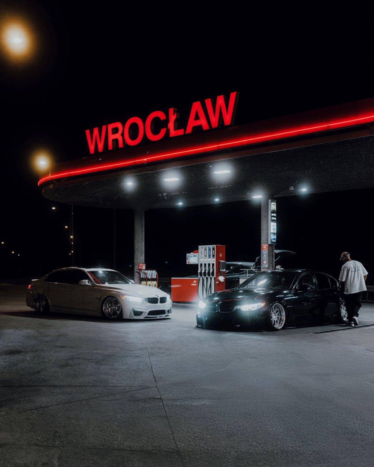 Two lowered BMWs under a gas station canopy with red Wrocław neon sign at night
