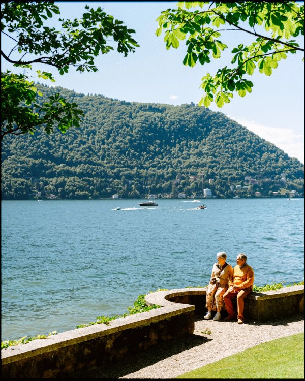 Older couple seated on a stone wall by Lake Como with mountain backdrop and leaves overhead
