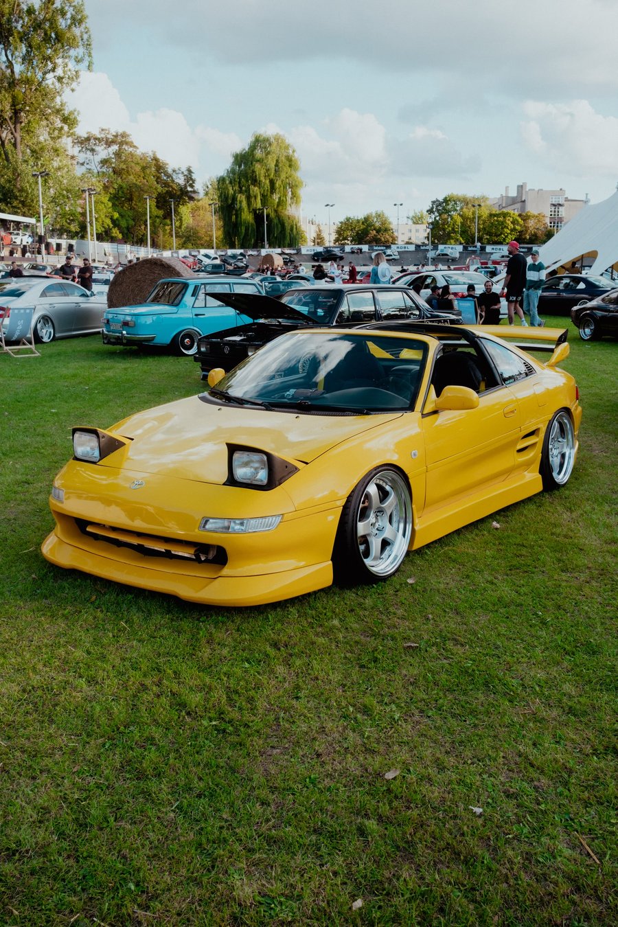 A yellow widebody Toyota MR2 on the grass infield, crowd and parked cars behind it