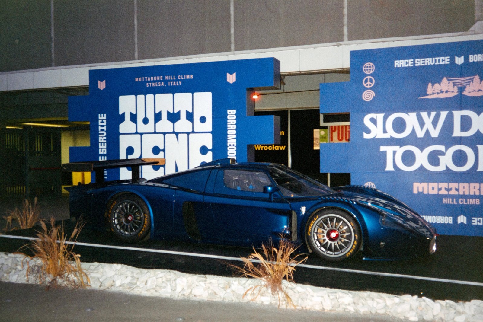 Maserati MC12 in dark blue parked in front of the Tutto Bene stand with blue typography