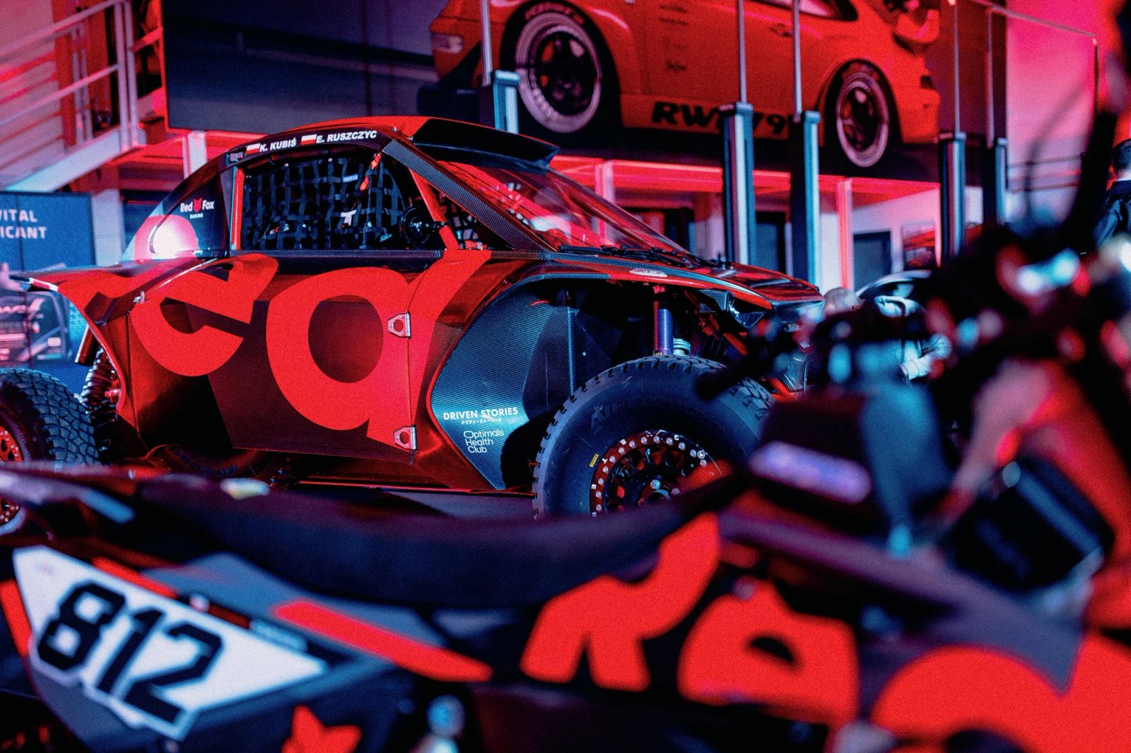 Black Taurus rally raid car with Red Fox livery and number 812 under red neon lights with a motorcycle beside it