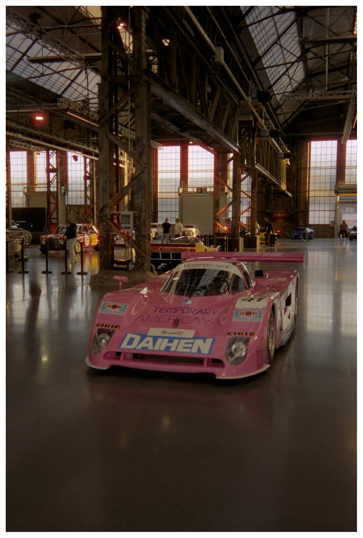 Pink Group C prototype with Daihen and Temporary Americaya sponsorship parked in an Areal Böhler hall with steel columns, industrial windows and a few people in the background