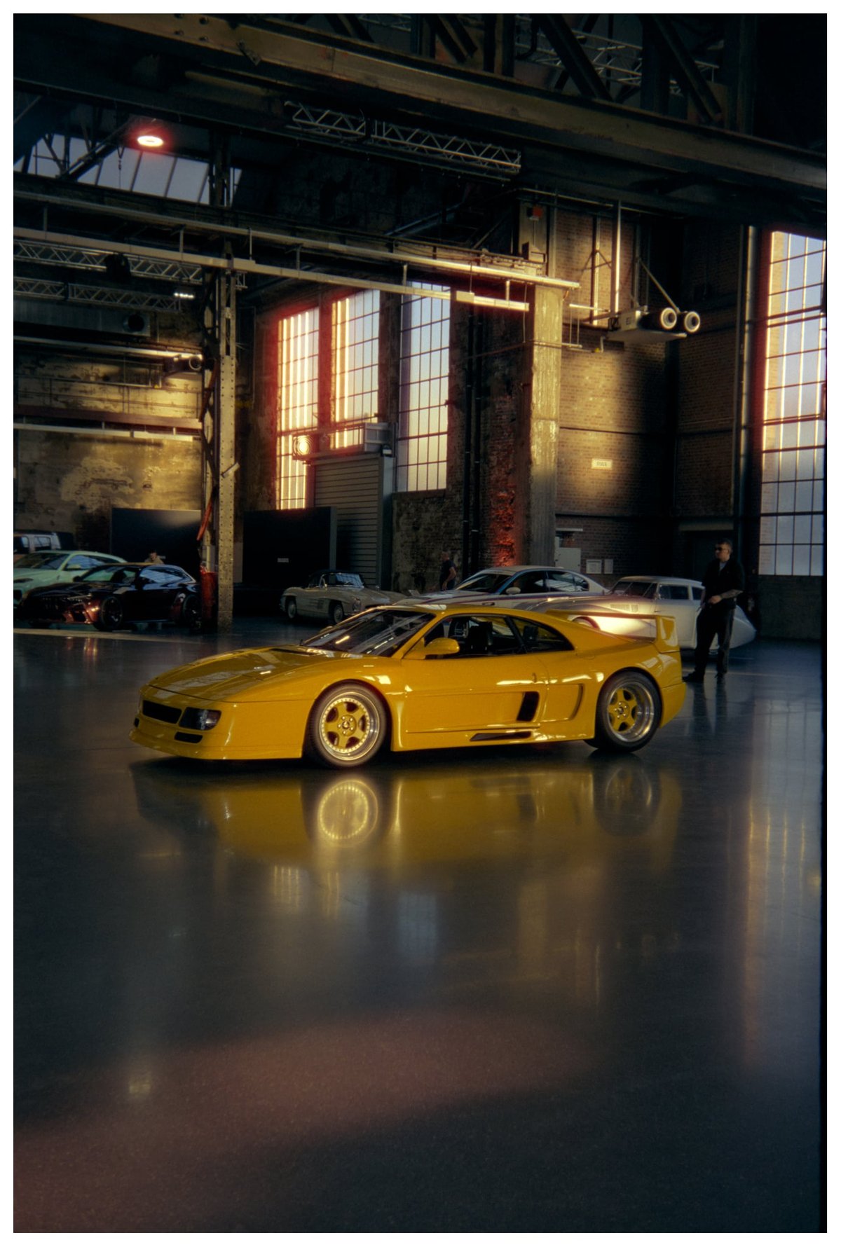 Yellow Ferrari 348 reworked by Koenig Specials as the F48 in side profile, parked on the reflective concrete floor of an Areal Böhler hall with golden window light behind it
