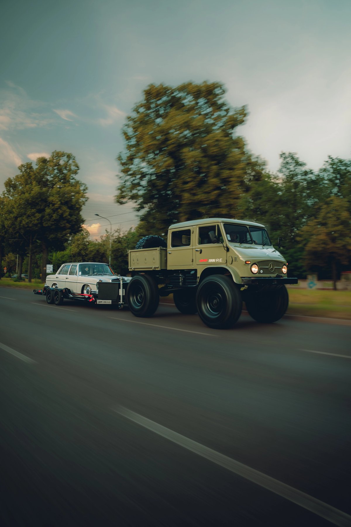 Unimog with W108 on trailer rolling shot from the side, trees blurred in motion