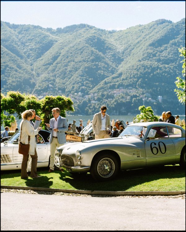 Silver vintage racing coupé number sixty with visitors examining it, Lake Como and Cernobbio mountains in the background