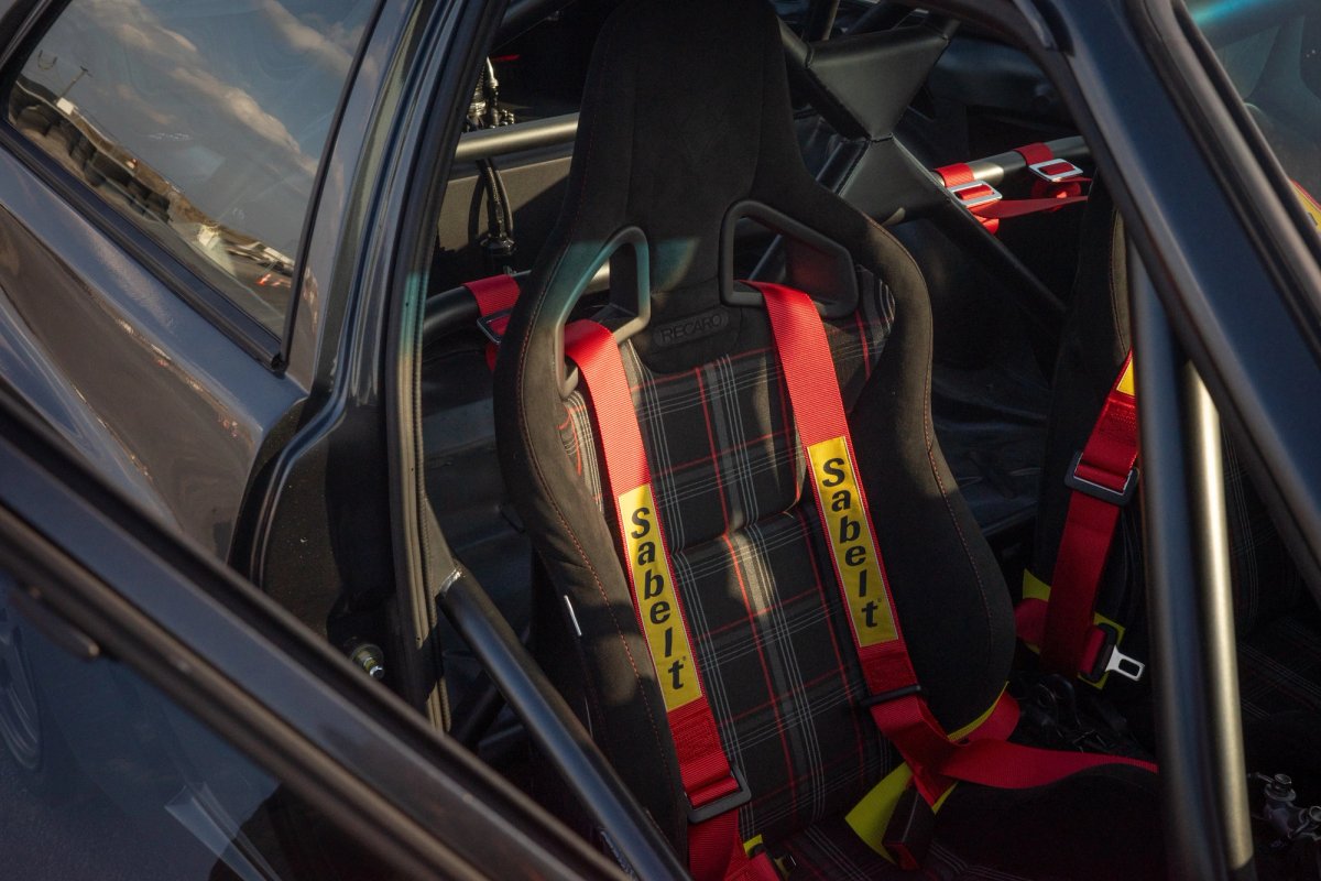 Recaro seat with tartan upholstery and red Sabelt six-point harness seen through the window