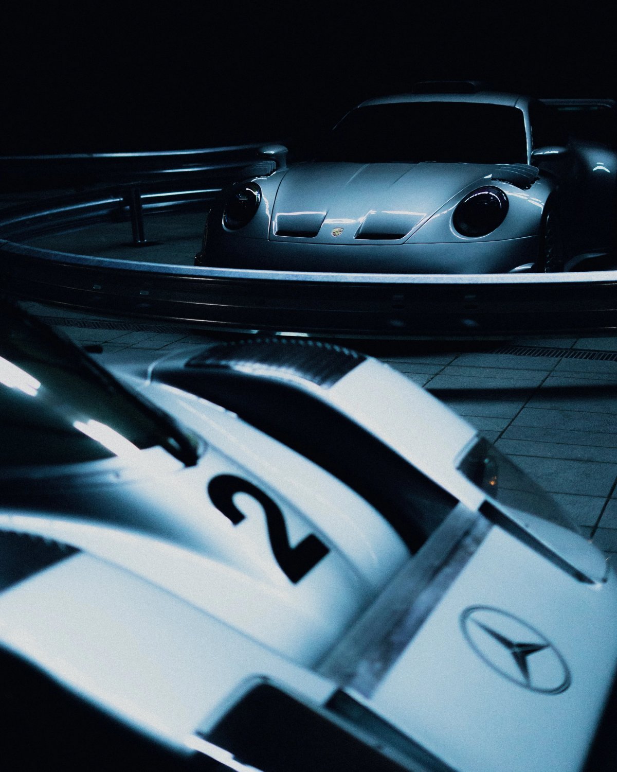 Silver Mercedes-Benz race car with the number two beside a Porsche 911 in cool indoor lighting