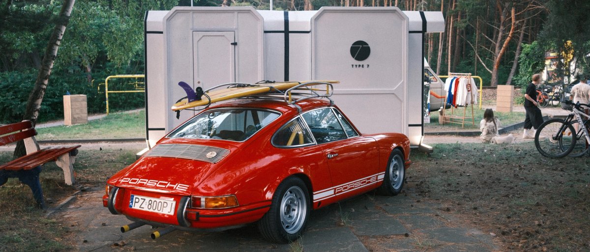 Red air-cooled Porsche 911 with two surfboards on a roof rack, parked in front of a white Type 7 camper pod at the Chałupy 6 campsite